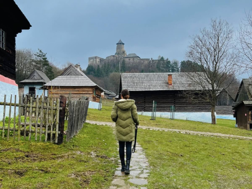 Žena kráča po cestičke pri tradičných chalúpkach, hrad v pozadí.